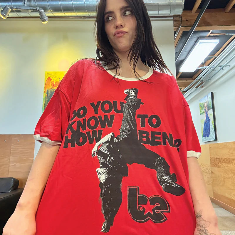 Billie Eilish wearing a red ringer tee with white trim, featuring a bold graphic of a breakdancer and the phrase “DO YOU KNOW HOW TO BEND?” with a stylized "be" logo. Statement streetwear merch with a playful twist and retro edge.