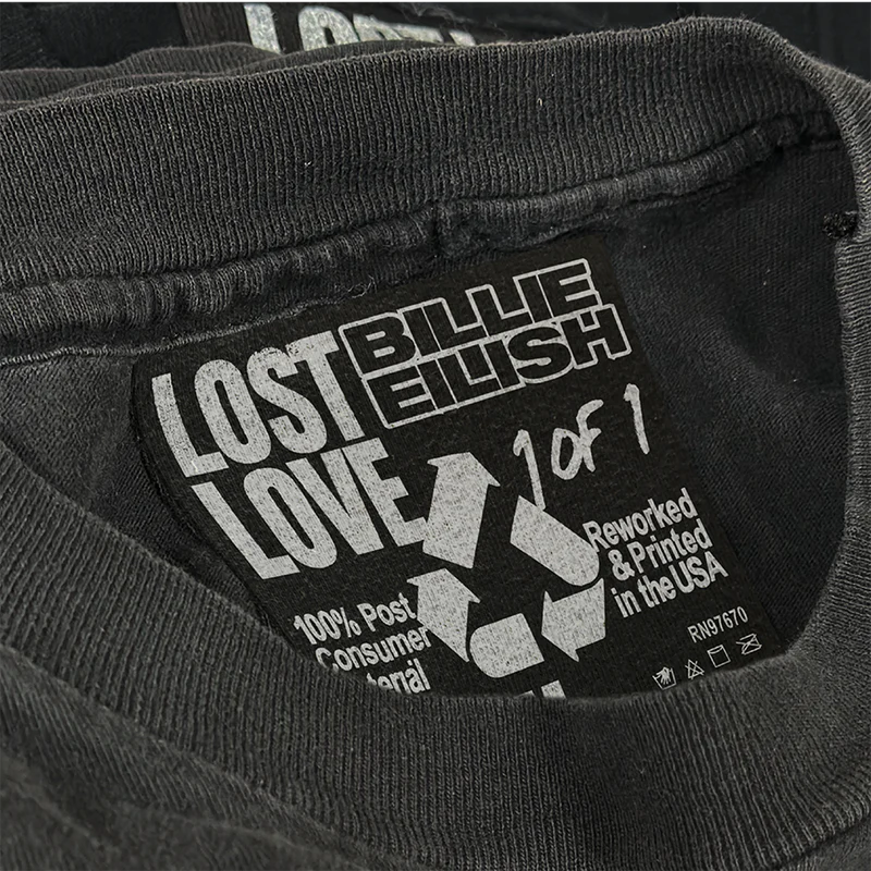 Close-up of the inner label on the Billie Eilish x Lost Love tee reading “1 of 1,” printed with a recycled symbol and text stating “100% post-consumer material” and “reworked & printed in the USA.” Unique, sustainable merch from the exclusive Billie Eilish collection.