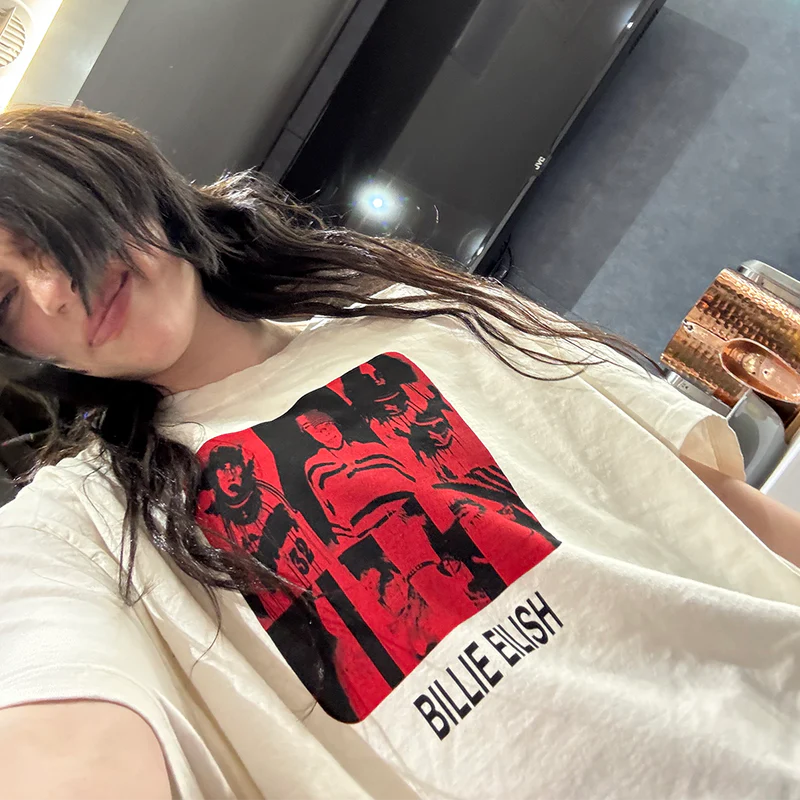 Billie Eilish smiling and wearing her Thermal BE Collage Tee, displaying the bold red-black collage artwork and black name text on a soft off-white T-shirt. Laid-back and expressive Billie Eilish merch for fans of her iconic aesthetic.