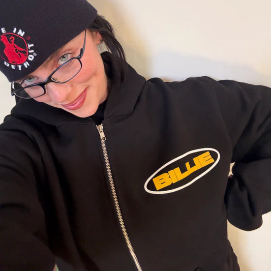 Billie Eilish smiling while wearing a black zip-up hoodie with a bold yellow "BILLIE" oval logo on the chest. Cozy and casual Billie Eilish hoodie, perfect for fans of her signature merch style.

