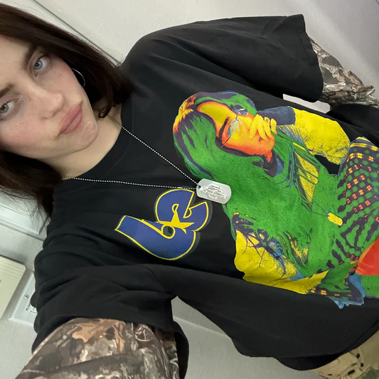 Billie Eilish wearing the "Hit Me Hard and Soft" camo layered long sleeve tee, featuring a bold, psychedelic portrait graphic in neon green, yellow, and orange tones. The black shirt includes camo-print sleeves and a “be” logo detail.

