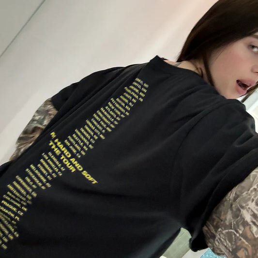 Back view of Billie Eilish modeling the "Hit Me Hard and Soft" tour tee, showing the full 2024 tour city lineup in yellow text. Layered camo sleeves and edgy styling create a unique streetwear look.

