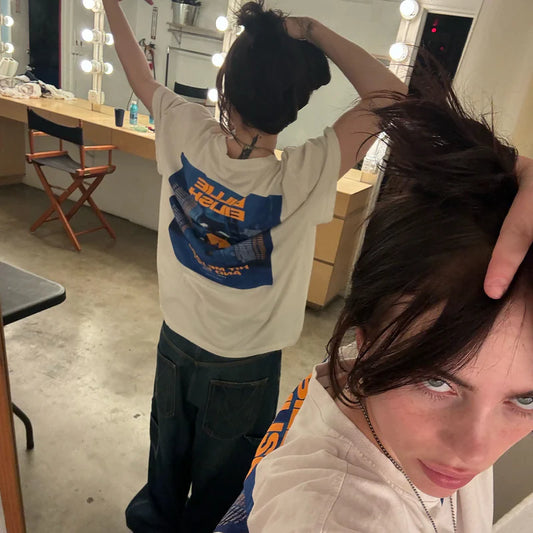 Billie Eilish taking a mirror selfie wearing the HIT ME HARD AND SOFT Admat Poster Tee, featuring the tour artwork and dates on the back in bold blue and orange. Behind-the-scenes fan-favorite look from her official 2024 merch line.

