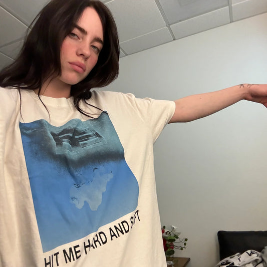 Billie Eilish wearing the “Hit Me Hard and Soft” white cover tee, featuring a blue-toned abstract graphic resembling underwater imagery and bold black text at the hem. A relaxed, artsy look tied to her latest album era.

