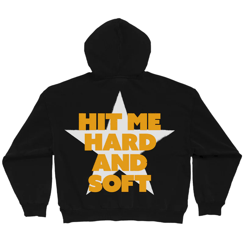 Flat-lay image of the Billie Eilish black hoodie with a white star and large yellow block text that says "HIT ME HARD AND SOFT." Official album-themed merch hoodie with oversized back graphic.

